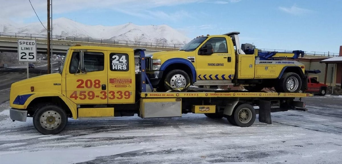 Yellow tow truck with flatbed carrying another yellow pickup truck, snowy mountains visible in background
